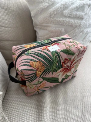 Blommig necessär från Emelo - Säljer en snygg necessär från Emelo med rosa botten och tropiskt blommönster i grönt, gult och vitt. Väskan har en svart dragkedja och praktiskt handtag på sidan. Perfekt för smink eller småprylar. Materialet är kraftigt tyg med beige insida. Innerfack. Mått ca 27x17x12cm