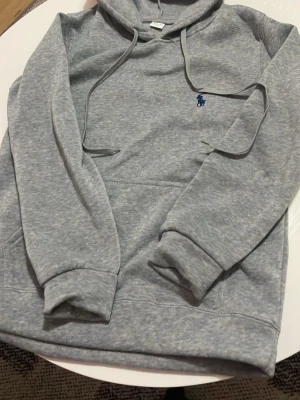 Grå hoodie från Polo Ralph Lauren - Snygg grå hoodie från Polo Ralph Lauren med klassisk blå logga broderad på bröstet. Hoodien har huva med dragsko, magficka och ribbade muddar vid ärmar och nederkant. Tillverkad i mjukt sweatshirtmaterial som är perfekt för chill dagar.