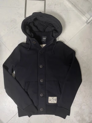 Mörkblå hoodie från Hollister - Snygg mörkblå hoodie från Hollister med knappar framtill, stor huva med dragsko och två fickor. Tjockt och mjukt material, perfekt för kyliga dagar. Hollister-logga på en patch vid fickan ger en clean och stilren look.