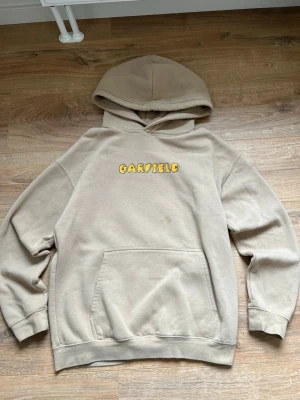 Beige Garfield hoodie med tryck - Säljer en beige hoodie med huva och känguruficka framtill. På bröstet finns ett gult Garfield-tryck i tecknad stil. Hoodien har ribbade muddar vid ärmar och nederkant. Perfekt för dig som gillar streetwear och vill ha något bekvämt och snyggt.