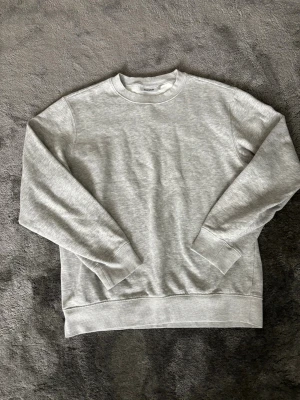 Grå sweatshirt från Weekday - En stilren grå sweatshirt från Weekday i storlek M. Tröjan har rund halsringning, ribbade muddar vid ärmslut och nederkant samt är gjord i mjukt bomullsmaterial. Perfekt för en avslappnad och clean look.