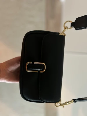 Svart axelväska Marc Jacobs - Oanvänd svart axelväska från Marc Jacobs med bred justerbar axelrem i beige och guld med logga. Väskan har guldfärgade metalldetaljer och en stilren, rektangulär form. 