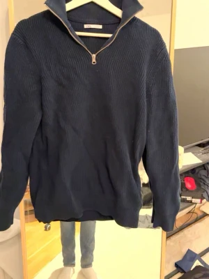 Mörkblå stickad tröja med halvzip - Mörkblå stickad tröja med hög krage och halv dragkedja framtill. Tröjan har ribbstickad struktur och långa ärmar.