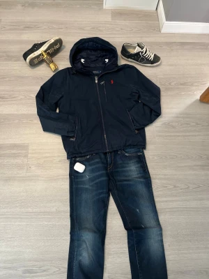 Mörkblå vindjacka från Polo Ralph Lauren - Snygg mörkblå vindjacka från Polo Ralph Lauren med huva och dragkedja framtill. Jackan har en bröstficka med dragkedja, två sidofickor och den klassiska röda loggan broderad på bröstet. Perfekt för våren  och enkel att matcha med jeans.