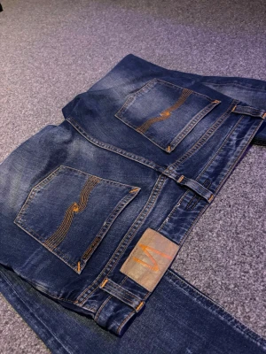 Nudie Jeans - Snygga blå slim fit jeans från Nudie Jeans med klassisk femficksdesign och orangea sömmar på bakfickorna. Jeansen har en lätt tvättad look och raka ben. Perfekta för en avslappnad och trendig stil.