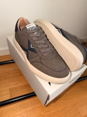 Alexander Smith - Snygga grå sneakers från Alexander Smith med marinblå detaljer och vit sula. Skorna har en klassisk låg siluett, grå skosnören och insida i beige läder. Perfekta för dig som gillar stilrena och moderna sneakers storlek 43