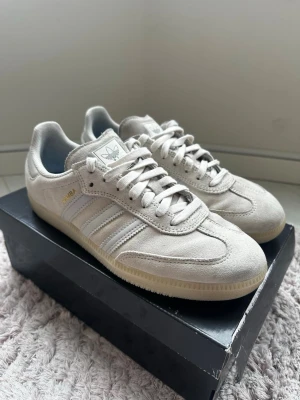 Adidas Samba OG beige mocka sneakers - Snygga Adidas Samba OG sneakers i beige mocka med klassiska vita ränder på sidorna och gummisula. Skorna har låg profil och ikonisk retrostil med guldfärgad Samba-text på sidan. Perfekta för dig som gillar streetwear och tidlös design.