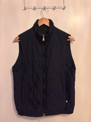 Oscar Jacobson mörkblå quiltad dunväst med dragkedja - Stilren mörkblå dunväst med quiltat mönster och hög krage. Västen har hel dragkedja framtill och två sidofickor. Perfekt att slänga över en hoodie eller tröja för extra värme. Materialet är lätt och smidigt, vilket gör den enkel att bära.