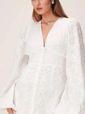 Vit kort adoore klänning - Supersnygg vit kort klänning med v-ringning, puffiga långärmar och dekorativa knappar framtill. Klänningen har ett subtilt blommigt mönster och är tillverkad i ett lätt tyg som ger en romantisk känsla. Perfekt för dig som vill sticka ut med en feminin och trendig look. 