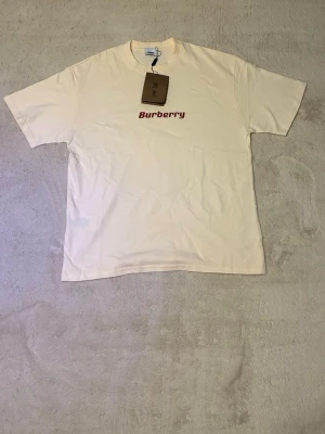 Beige Burberry t-shirt med tryck - Cool beige t-shirt från Burberry med röd logga framtill och ett stort rött spöke-tryck på ryggen. Klassisk passform och rund halsringning. Perfekt för dig som vill sticka ut med en unik design och streetwear-vibe.