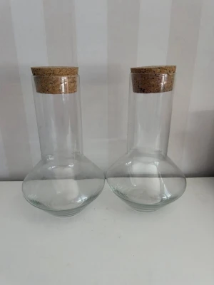 Två glasflaskor med korklock - Säljer två stilrena glasflaskor med bred botten och smal hals, perfekta för vatten, juice eller dekoration. Flaskorna är genomskinliga och har naturliga korklock som ger en modern och enkel look. Passar bra i köket eller som inredningsdetalj.