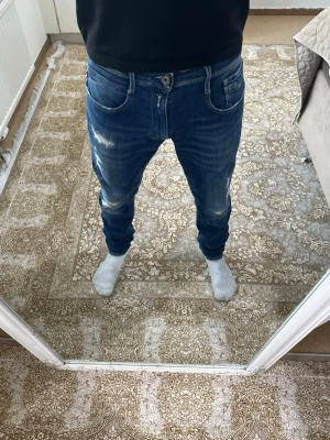 Blå slitna jeans från Replay - Säljer ett par blå jeans från replay med snygga slitningar och tvättade detaljer på framsidan. Jeansen har fem fickor, normal midja och smal passform. Klassisk blå färg och tillverkade i stretchigt denimtyg för extra komfort. Hör av er priset kan diskuteras.