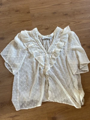 Vit blus med volang från Zara - Söt vit blus från Zara med volangdetaljer längs knäppningen och prickigt strukturmönster. Blusen är gjord i ett tunt, transparent tyg och har trekvartsärm. Perfekt för en trendig och luftig look. Storlek M men passar en S/Xs