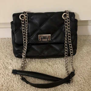 Svart quiltad axelväska med kedja - Snygg svart axelväska i quiltad design med silverfärgad kedjerem och vridlås framtill. Väskan har en mjuk, puffig form och detaljer i metall som ger en trendig look. Från stradivarius 