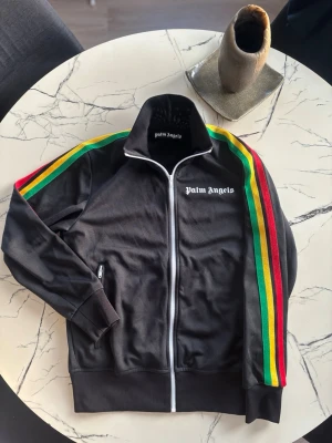 Palm Angels svart zip-tröja (S) - Palm Angels svart zip-tröja med vita detaljer och logga på bröstet. Färgglada ränder i rött, gult och grönt längs ärmarna. Tröjan har hög krage, dragkedja och sidofickor. Perfekt för en cool streetstyle-look.