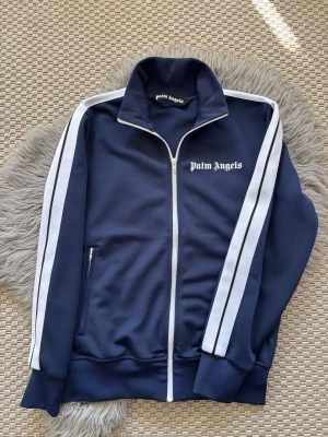 Palm Angels marinblå zip-tröja (M) - Palm Angels zip-tröja i marinblått med vita detaljer och logga på bröstet. Tröjan har hög krage, vita dragkedjor och vita ränder längs ärmarna. Perfekt för en sportig och avslappnad look. Materialet är mjukt och stretchigt.