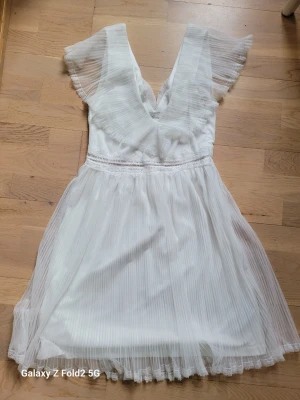 Vit plisserad klänning med spets - Superfin vit kortklänning från NLY Trend med plisserat tyg, v-ringning och spetsdetaljer vid midjan och ärmarna. Klänningen har korta, transparenta ärmar och en flowy kjol som ger en romantisk vibe. Perfekt för sommarens alla tillfällen!