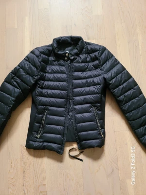 Cardigan dunjacka från Zara Woman - Snygg svart dunjacka från Zara Woman med dragkedja och tryckknappar framtill. Jackan har två fickor med dragkedja och en hög krage. Perfekt för kalla vinterdagar och har en stilren, figurnära passform.