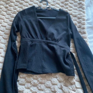 Svart omlottopp med knytband - Snygg svart omlottopp med långa ärmar och knytband i midjan. Toppen har en djup v-ringning och är gjord i ett lätt, strukturerat tyg som ger en stilren look. Perfekt att styla med jeans eller kjol för en trendig outfit.