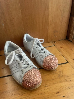 Vita sneakers med roséguld glitter - Unika vita Adidas sneakers med klassiska tre ränder på sidan och snörning. Tåpartiet är täckt av glittrande roséguld dekor som ger skorna en personlig och trendig touch. Perfekta för dig som vill sticka ut med din stil.