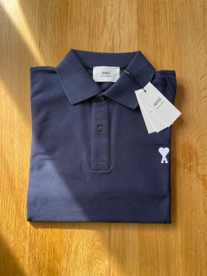 Mörkblå pikétröja från Ami - Snygg mörkblå pikétröja från Ami med klassisk krage och korta ärmar. Tröjan har knäppning framtill och Ami-logga med hjärta broderad på bröstet. Perfekt till jeans eller chinos för en clean look.