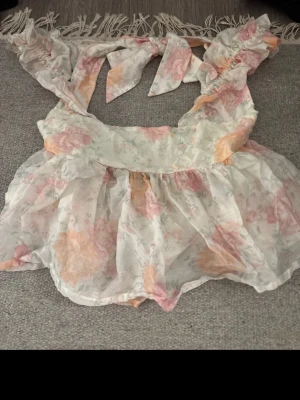 Blommig volangblus med knytband - Säljer en romantisk blommig blus med volanger och knytband i ljusa pastellfärger. Blusen är ärmlös och har en luftig, transparent look med stora blommor i rosa, orange och beige. Perfekt för dig som gillar en feminin och trendig stil. Passar mig som brukar bära s/xs