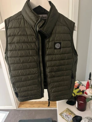 Stone Island junior dunväst - Snygg olivgrön tunn dunväst från Stone Island med quiltad design och hög krage. Västen har dragkedja framtill, två sidofickor med dragkedja och den klassiska Stone Island-loggan på bröstet. Tillverkad i lätt och värmande material, perfekt för lager-på-lager.