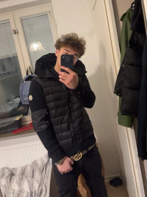 Moncler Cardigan  - Säljer en svart pufferjacka från Moncler med huva och diskret logga på ärmen. Jackan har quiltad design på kroppen och släta svarta ärmar. Perfekt för dig som vill ha en stilren och trendig vinterjacka. NFC funkar