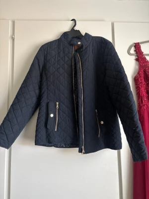 Mörkblå quiltad jacka med dragkedja - Snygg mörkblå quiltad jacka med ståkrage och dragkedja framtill. Jackan har två fickor med dragkedja och knappdetaljer. Perfekt för dig som vill ha en stilren och enkel jacka till vardags. Materialet är lätt vadderat för extra komfort.