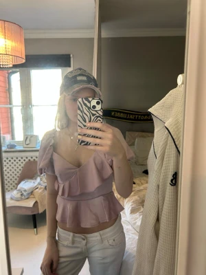 Ljuslila volangblus med puffärm - Supersöt ljuslila/rosa blus med volanger och puffärmar. Toppen har v-ringning och är figurnära i midjan, vilket ger en feminin siluett. Perfekt för dig som vill ha något gulligt och trendigt. Materialet känns mjukt och lätt.