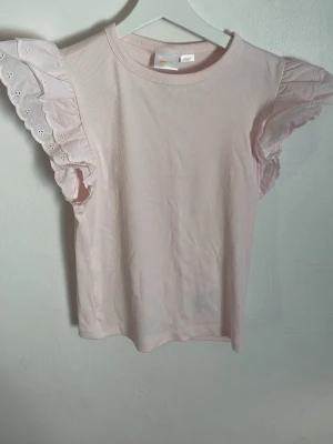 Ljusrosa topp med volangärmar - Supersöt ljusrosa topp med ribbad struktur och breda volangärmar i spets. Perfekt för dig som gillar romantiska detaljer och vill ha något extra till din outfit. Toppen har rund halsringning och är i mjuk bomull.
