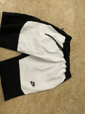 Svarta och vita Nike shorts med snörning - Säljer ett par snygga svartvita shorts från Nike med elastisk midja och snörning. Shortsen har en sportig look med kontrastpaneler och Nike-logga på benet. Perfekta för träning eller chill dagar.