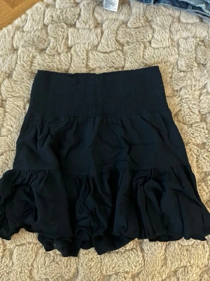 Svart volangkjol med resårmidja - Säljer en svart kortkjol med bred resår i midjan och flera lager volanger. Kjolen har en luftig och flowy känsla, perfekt för att styla med en enkel topp. Materialet känns lätt och bekvämt.