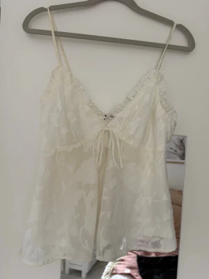 Vit linnetopp med volang och knyt - Söt vit linnetopp med tunna axelband, volangdetaljer vid urringningen och knyt framtill. Toppen har ett subtilt blommigt mönster i tyget och är lätt och luftig, perfekt för sommaren med ett par linnebyxor eller kjol