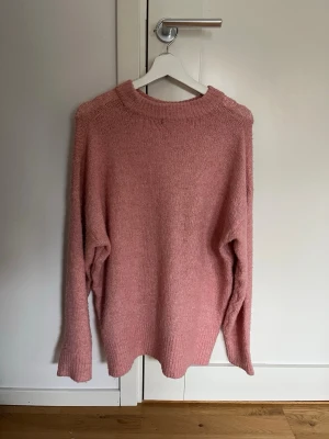 Rosa stickad tröja med rund hals - Mysig rosa stickad tröja med rund hals och långa ärmar. Tröjan har en enkel, rak passform och ribbstickade muddar vid halsen. Perfekt för lager på lager och en avslappnad stil.