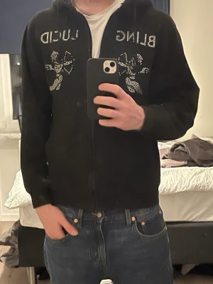 Svart hoodie med blingdetaljer - Säljer en svart hoodie från bling lucid med blingade stenar och texten 'bling lucid' på bröstet. Dragkedja framtill och huva. Perfekt för dig som gillar statement-plagg och vill sticka ut lite extra.