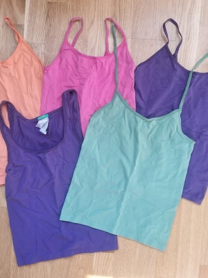 Färgglada linnetoppar i fempack - Fem olika linnetoppar i färgerna lila, grön, rosa, orange och mörklila. Alla har smala axelband och är tillverkade i mjuk bomull, perfekta för lager på lager eller varma dagar. Säljs som ett paket för dig som vill ha variation i garderoben. Obs! Det banden på det gröna linnet måstensys fast.
