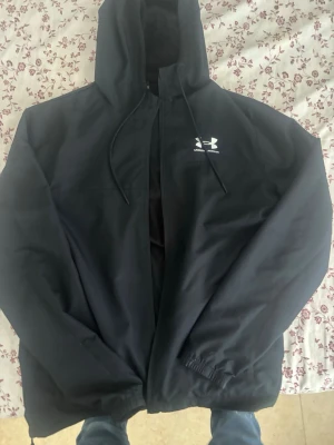 Svart vindjacka från Under Armour - Snygg svart vindjacka från Under Armour med huva och dragsko. Jackan har dragkedja framtill och en vit logga på bröstet. Tillverkad i lätt och vindtätt material, perfekt för blåsiga dagar.
