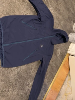 Mörkblå jacka från Haglöfs - Snygg mörkblå vindjacka från Haglöfs med huva och två dragkedjeförsedda bröstfickor. Jackan är använd bara 2 gånger 