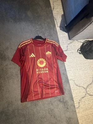 Röd AS Roma matchtröja Adidas - Snygg röd matchtröja från AS Roma tröja med gula detaljer och ränder. Adidas-logga på bröstet, klubbmärke och sponsortryck framtill. Nummer 26 och namnet 'Dahl' på ryggen. Tillverkad i lätt och ventilerande material, perfekt för fotbollsträning.