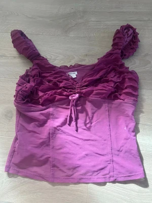 Lila volangtopp från Kimchi Blue - Supersöt lila topp från Kimchi Blue med volanger över bysten och axelbanden. Toppen har en snygg knytdetalj framtill och är ärmlös. Perfekt för dig som vill sticka ut med en färgglad och unik look.