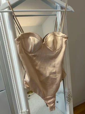 Beige satinbody med bygelkupor - Snygg beige body i satinliknande material med bygelkupor och dubbla tunna axelband. Ger en lyxig känsla och har en figurnära passform som framhäver siluetten. Perfekt att styla under en kavaj eller till jeans för en trendig look.