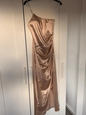 BALKLÄNNING /bröllopsklänning - Snygg långklänning i champagnefärg med glansigt material och en axelbandslös design. Klänningen har en hög slits framtill och draperad effekt längs sidan. Perfekt för dig som vill sticka ut med en elegant look.