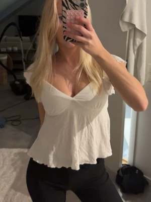 Vit v-ringad blus med volang - Säljer en söt vit blus med v-ringning och volangkant. Toppen har korta ärmar och en lätt croppad passform som ger en avslappnad men feminin look. Perfekt att styla med jeans eller kjol för en trendig outfit.