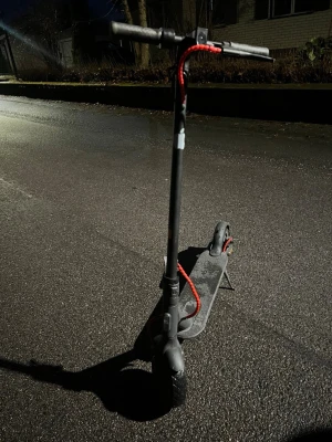 Svart elsparkcykel med röda detaljer - Säljer en svart elsparkcykel med röda kablar och gråa handtag. Den har en robust ram i metall, stora hjul och stödben. Perfekt för att ta sig fram snabbt i stan eller till skolan. Laddningsuttag och broms bak syns tydligt.