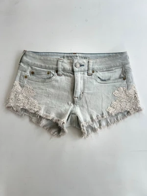Ljusblå jeansshorts med spetsdetaljer - Snygga ljusblå jeansshorts ifrån American Eagle med fransig kant och vita spetsapplikationer på sidorna. Klassisk femficksmodell med knapp och dragkedja framtill. Perfekta för sommaren och ger en avslappnad men söt look.