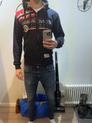 Mörkblå Geographical Norway hoodie - Snygg mörkblå hoodie från Geographical Norway med dragkedja, huva och stora tryckta loggor samt norska flaggan på bröstet. Röd och vit detalj upptill, ribbade muddar och fickor framtill. Perfekt för en sportig och avslappnad stil. Perfekt till det varmare vädret som kommer nu. 