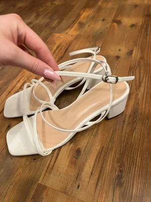 Vita sandaler med remmar och klack - Stilrena vita sandaler med tunna remmar och fyrkantig tå. Sandalerna har en låg, bred klack och justerbart spänne runt ankeln. Klacken är 5cm, aldrig använda. Perfekta till studenten eller sommaren! 