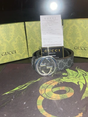Svart GG-mönstrat bälte från GG - Snyggt svart bälte från GG med klassiskt GG-mönster och stor silverfärgad logga som spänne. Bältet har en modern design och är tillverkat i skinn med detaljer i syntet. Perfekt accessoar för att lyfta din outfit.