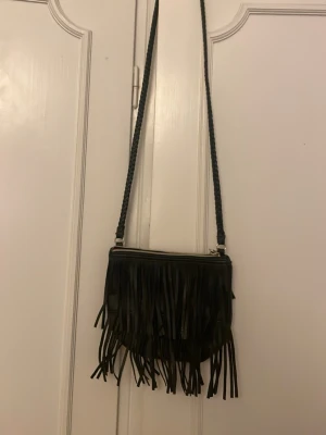 Svart axelväska med fransar - Cool svart axelväska i fejkläder med långa fransar framtill och flätat axelband. Väskan har dragkedja upptill och en stilren, bohemisk vibe som sticker ut. Perfekt för dig som vill addera lite edge till din outfit.
